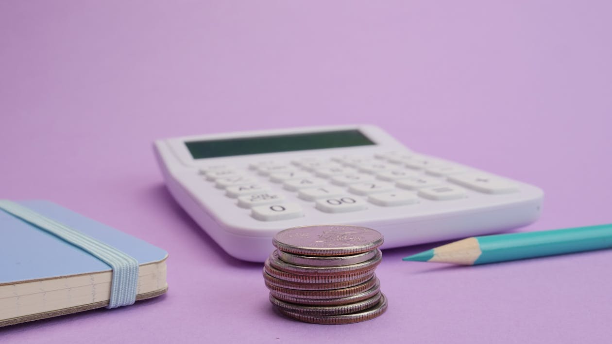 Calculadora branca, moedas empilhadas e caderneta azul sobre fundo lilás representando como montar um orçamento mensal