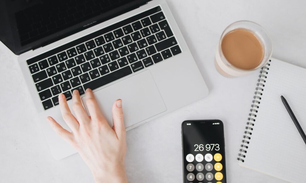 Mãos no notebook ao lado de calculadora e celular, fazendo a conta de CLT ou PJ