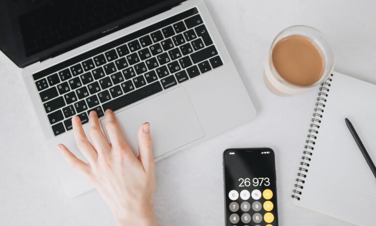 Mãos no notebook ao lado de calculadora e celular, fazendo a conta de CLT ou PJ