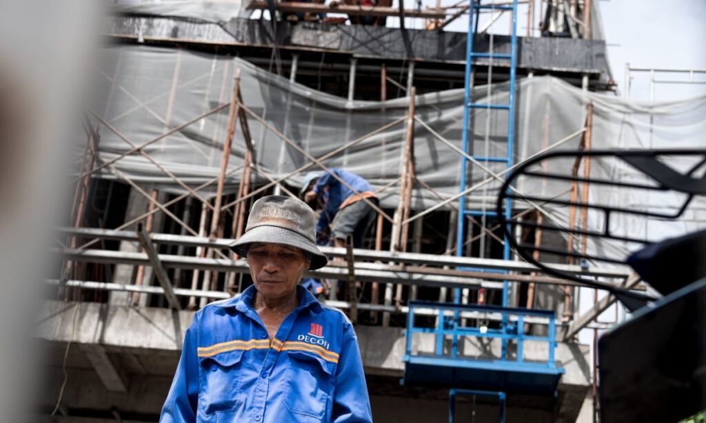 Trabalhador da construção civil de uniforme e chapéu em frente a uma obra, público do salário mínimo 2027